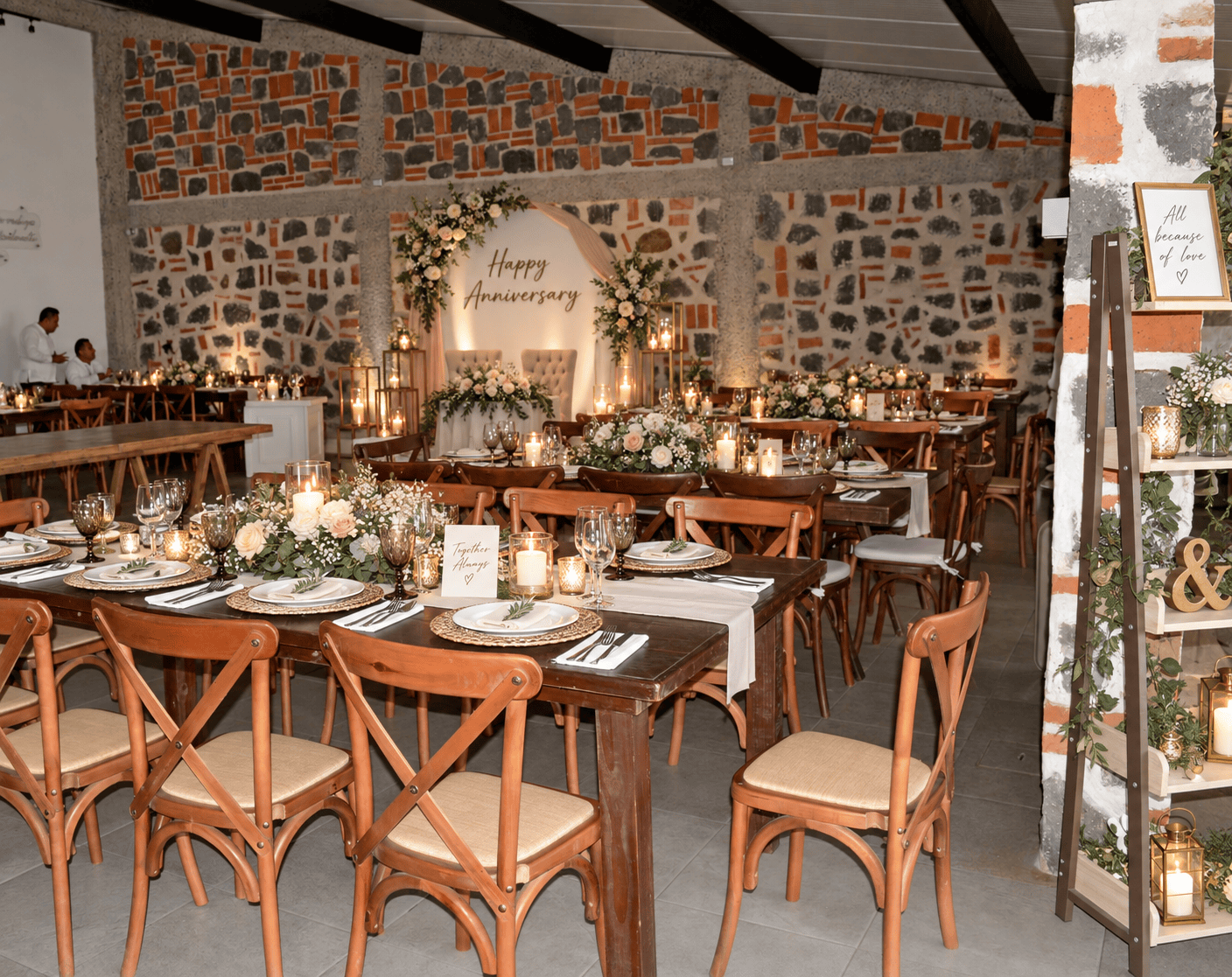 Salón de muros de piedra ambientado para aniversario con letrero Happy Anniversary, mesas de madera, candelas y arreglos florales