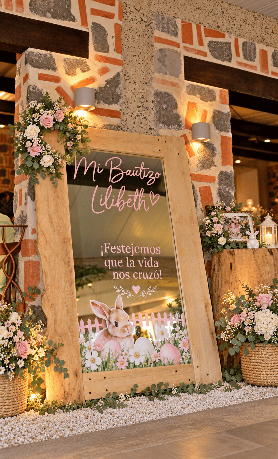 Espejo de bienvenida decorado para un bautizo con letras rosas, flores rosadas y blancas sobre muro de piedra