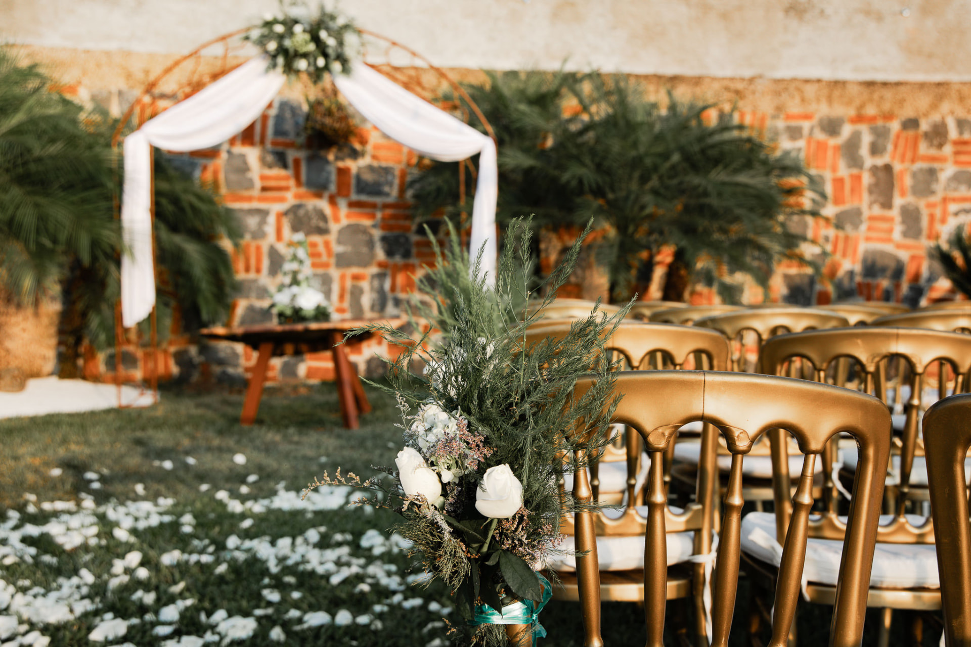 Jardín listo para ceremonia de boda con sillas doradas, arco de metal con telas blancas, flores y pétalos sobre el pasto