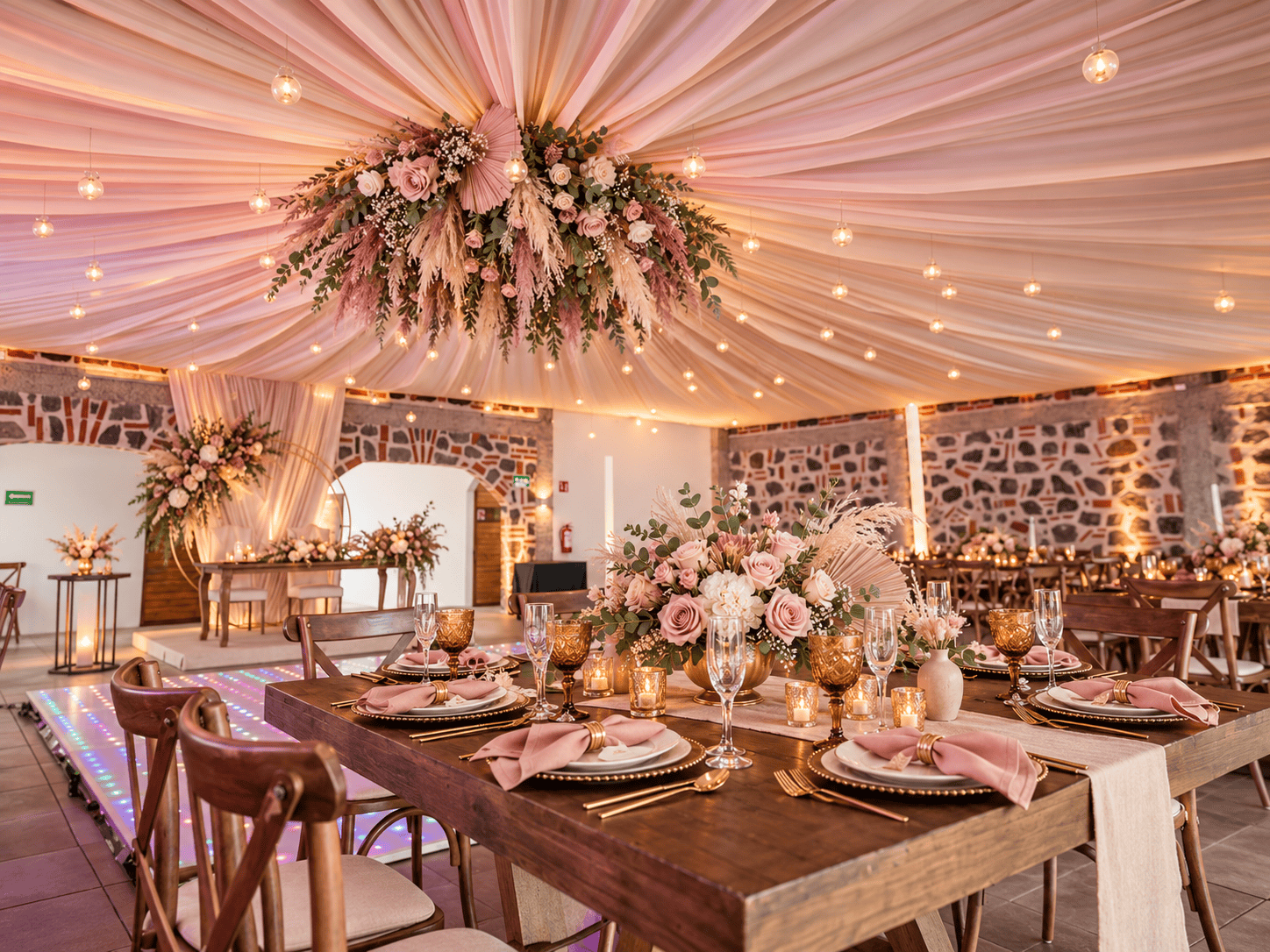Salón con toldo de tela rosa y arreglos colgantes, mesas de madera con flores rosadas, copas doradas y pista iluminada