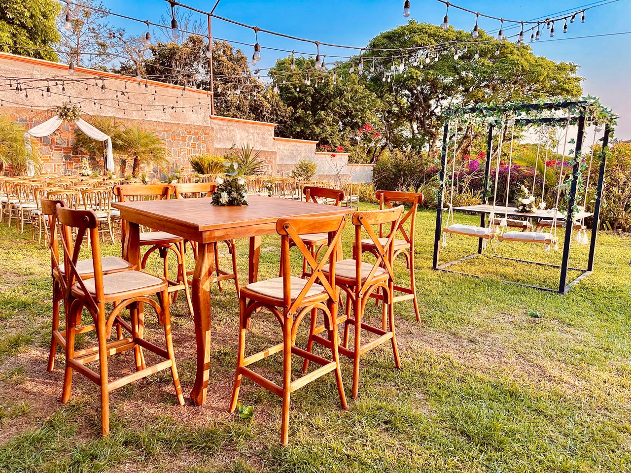 Jardín al aire libre con mesa alta de madera y bancos, pérgola negra con columpios y flores blancas, guirnaldas de luces y muro de piedra entre palmeras y arbustos en flor
