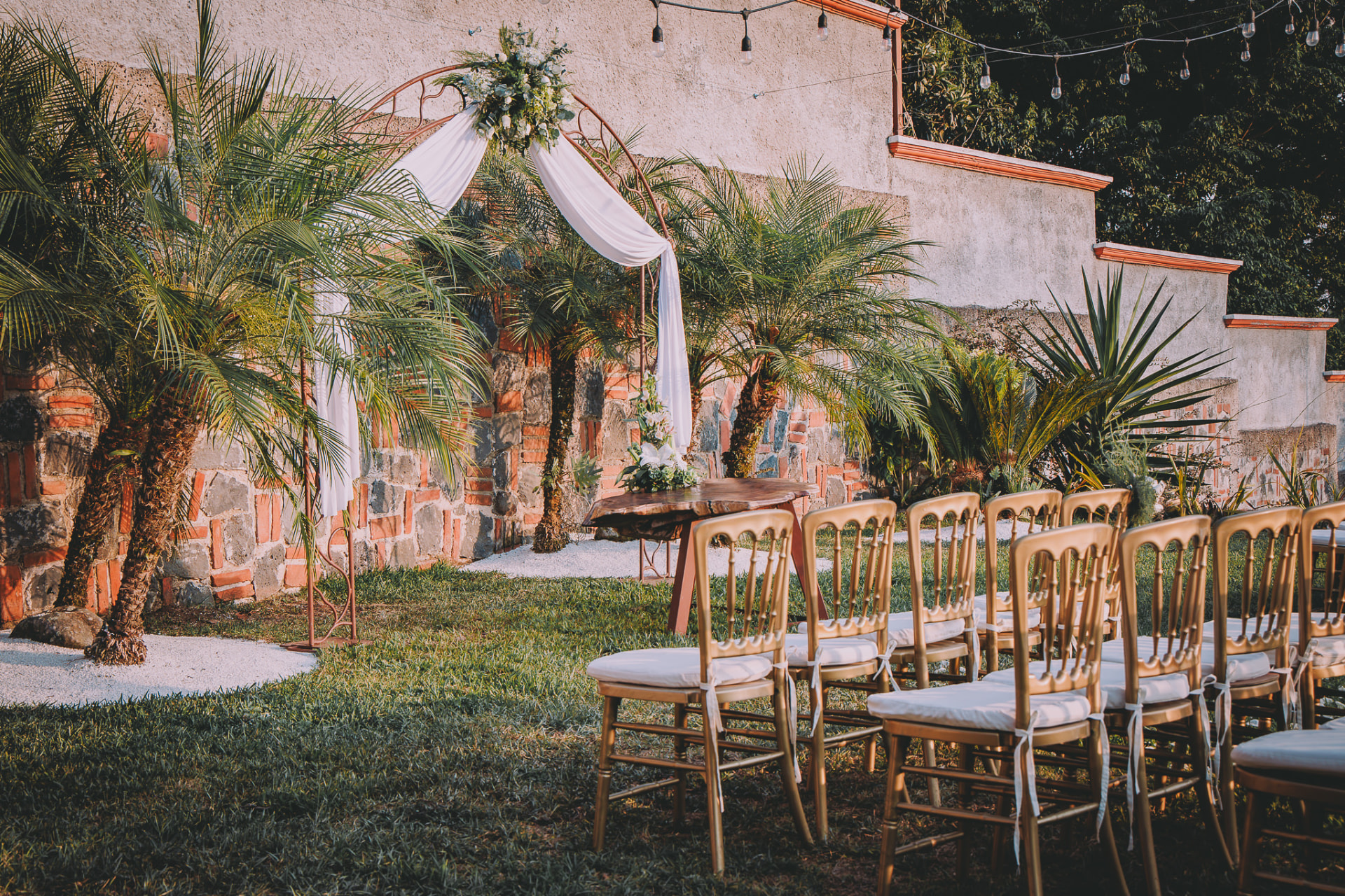 Ceremonia al aire libre con arco floral, sillas doradas y palmeras en los jardines de Quinta Los Mulatos